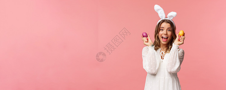 跳舞的兔子假期 春天和聚会的概念 兴奋迷人的年轻女性穿着兔耳和白色派对礼服庆祝复活节的肖像 用两个彩蛋跳舞 粉红色背景 成人 女朋友背景