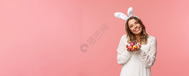 拿着小旗兔子假期 春天和聚会的概念 身着白色裙子和兔耳的肖像温柔浪漫的金发女孩 倾斜的头很可爱 微笑着开心地拿着彩蛋 庆祝复活节 粉红色的背背景