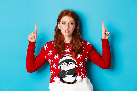 圣诞快乐 怀疑和没有被嘲笑的红发女孩举起手指 用不情愿的脸脸显示徽标 站在蓝背景上 假期 漂亮的背景图片