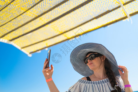 夏季海滩假期 沙滩上 一位脸上长着雀斑的红发少女头戴大檐帽 披着轻便的斗篷 遮住烈日 在遮阳棚下自拍 女孩微笑着享受阳光明媚的一背景