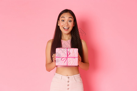 节日 庆典和生活方式概念 欢乐的亚洲女孩庆祝生日会收到包着的礼物 想知道现在里面是什么 在粉红背景上站得开心起来背景图片