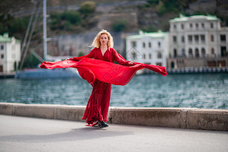 柳岩红裙飞舞身着红裙的女子 长着丝绸长翅膀的时装模特 身着飘逸的裙子 在堤岸上飞舞着布料 水 细胞背景