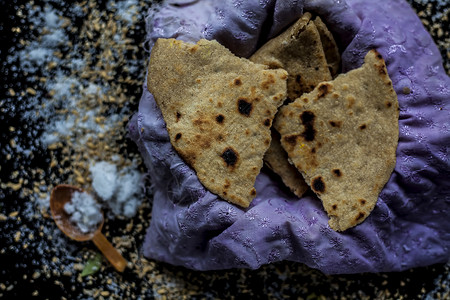 特写镜头：黑色木质表面上的圆形面包 Bhakri 以及容器中的一些生全麦和盐图片