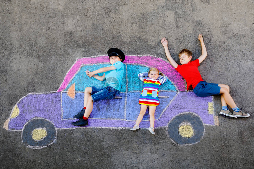 三个小孩两个学童男孩和蹒跚学步的女孩在沥青上用彩色粉笔画汽车画很开心兄弟姐妹一起在地上画图片