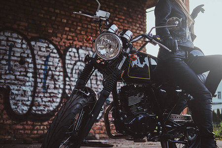 穿着摩托服装的时髦感女人正在她自行车和涂鸦墙旁边为图片