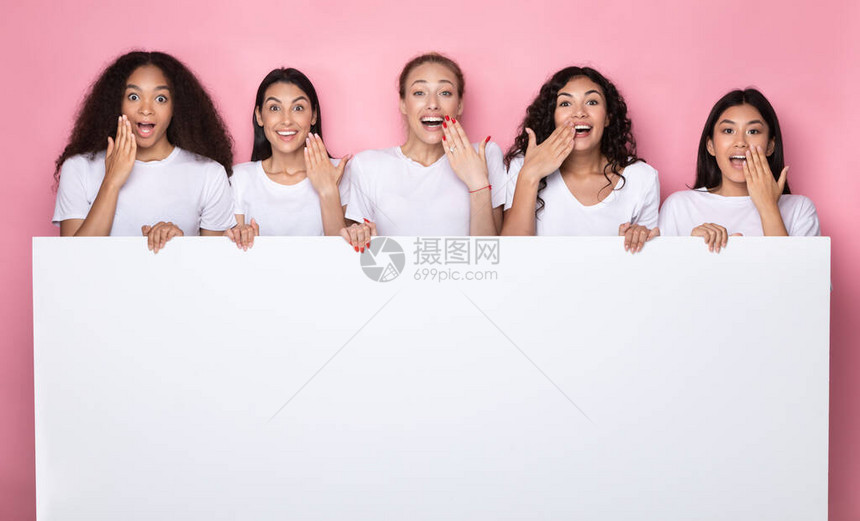 兴奋的多民族女士拿着空白纸海报与复制空间广告你的文字站在粉红色的工作室背景图片