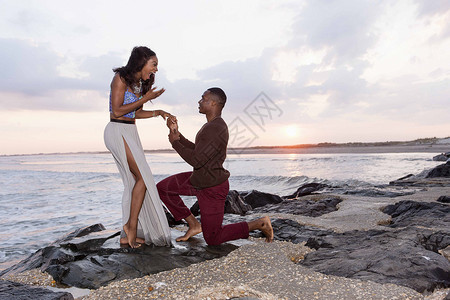 中年男子跪在海边的岩石上向年轻女子求婚图片