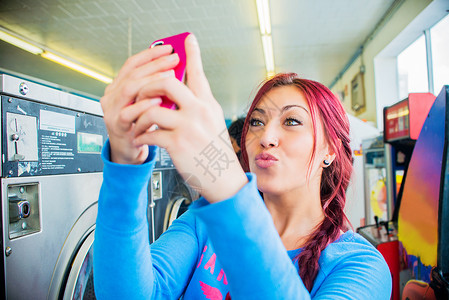 年轻女子在自助洗衣店用智能手机自拍图片