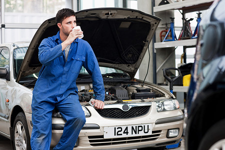 汽车发动力年轻的机修工在车上喝咖啡背景