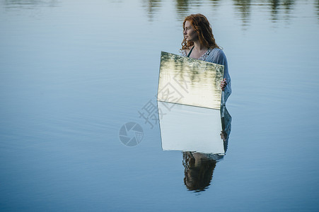 年轻女子站在湖中举着镜子水中的涟漪在镜子中反射图片