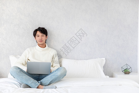 居家男性坐在床上用电脑查阅资料青年男性坐在床上使用电脑背景