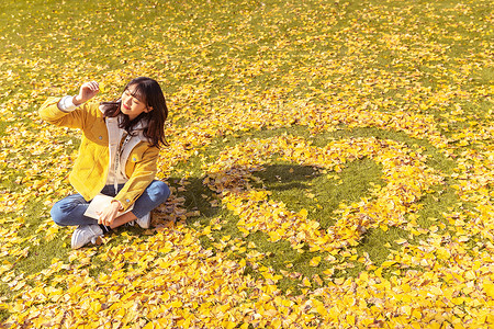 金色爱心光效坐在铺满银杏叶的草坪上看书的女孩背景