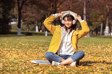 可爱女孩读书坐在铺满银杏叶的草坪上听音乐的女孩背景