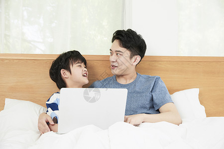 年轻爸爸陪儿子在床上使用笔记本电脑图片