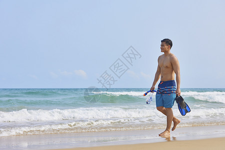潜水脚蹼男青年拿着潜水装备在海边行走背景