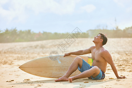 沙滩冲浪男孩年轻男士坐沙滩上拿着冲浪板背景