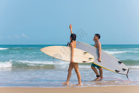 沙滩裤青年情侣拿着冲浪板海边沙滩上行走背景