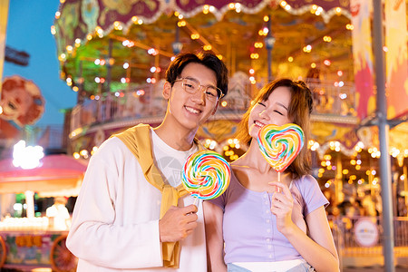 爱在七夕情人节拿着棒棒糖的年轻情侣在游乐园约会背景