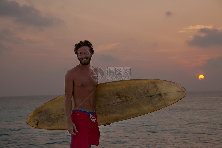 日落时在海滩上拿着冲浪板的男性图片