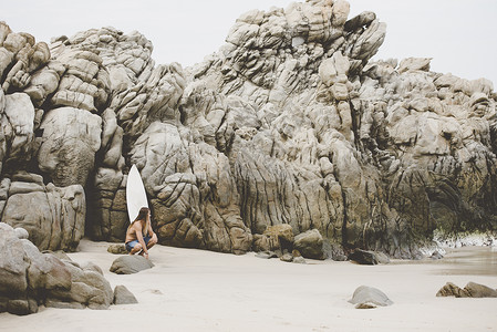 坐在海边岩石上的男人手拿冲浪板图片