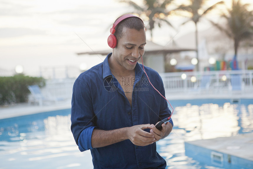 在游泳池边戴着耳机使用智能手机的中年男性图片