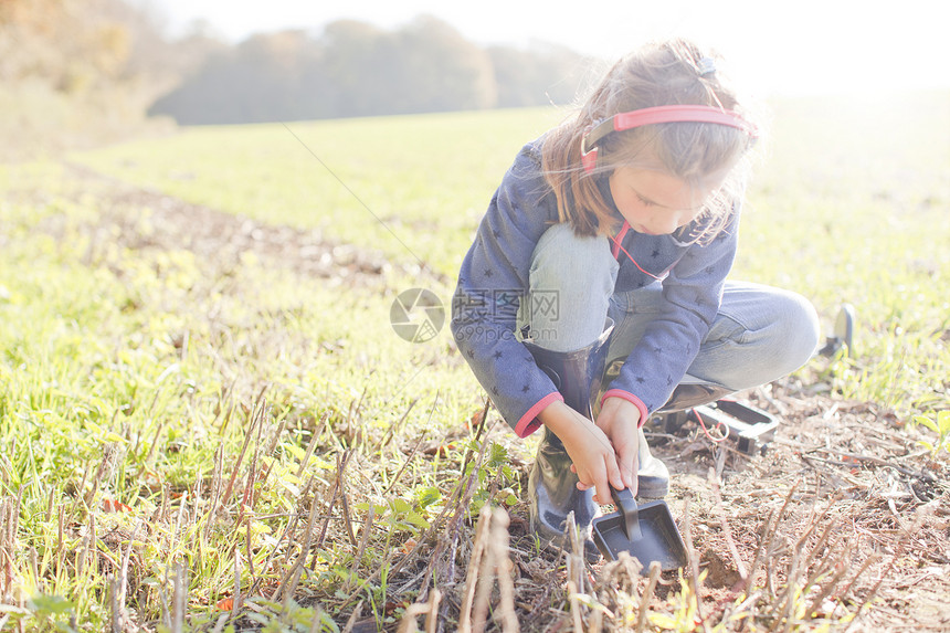 有金属探测器的女孩蹲在地上用铲子挖图片