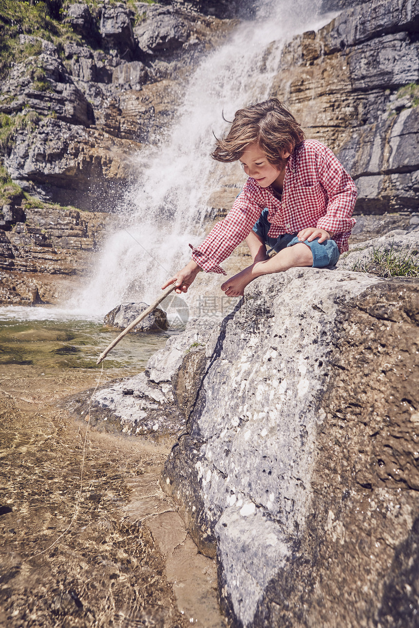 年轻坐在瀑布边的岩石上握着棍子看水图片
