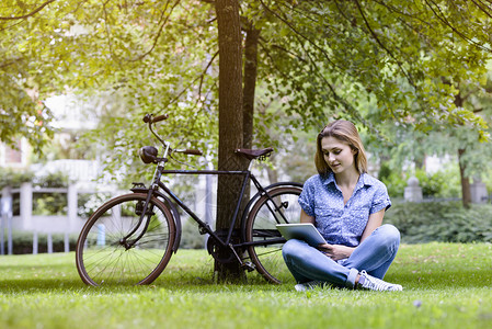 坐在自行车旁边的草地上用数字平板交叉脚的年轻妇女图片