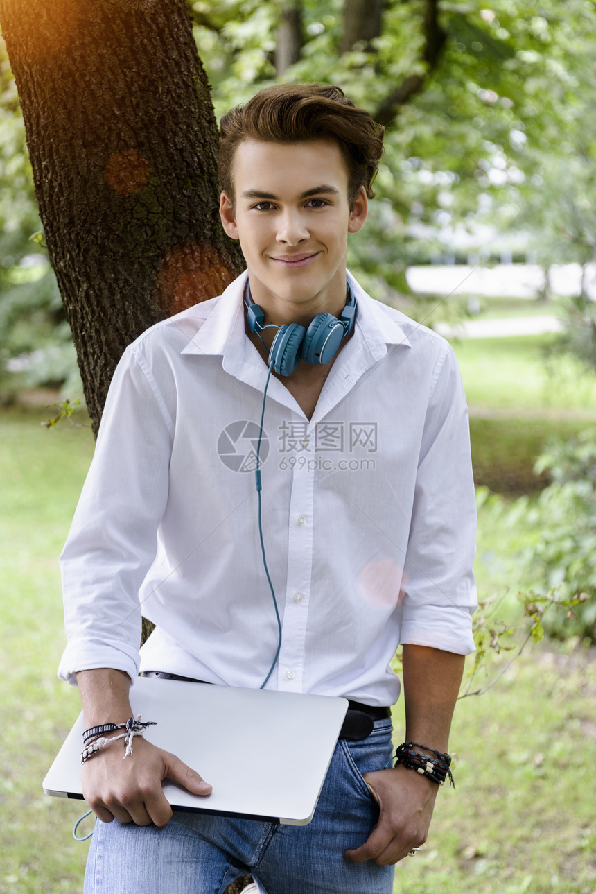 拿着耳机的年轻人靠在树上手放口袋里拿着笔记本电脑看着镜头微笑图片
