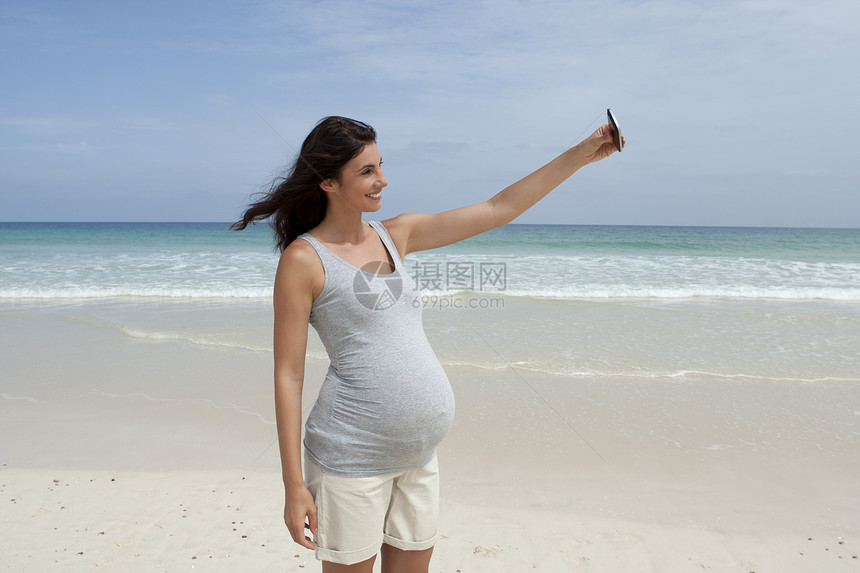 怀孕的中年成妇在非洲佛得角海滩使用智能手机自拍图片