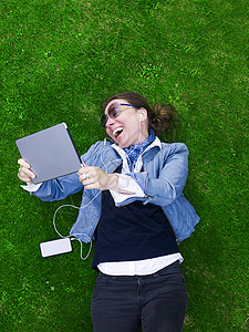 坐在草地上欢笑的成人却拿着数字平板电脑的高视线图片