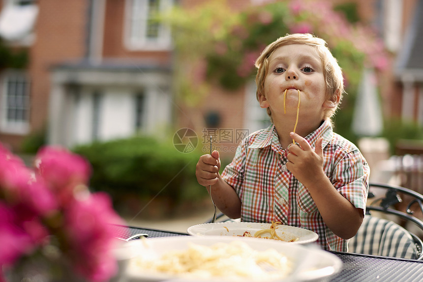 坐在花园桌上的男孩吸着意大利面看摄像机图片