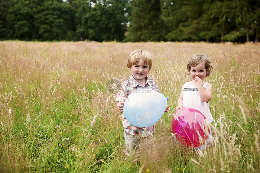 草地上拿着气球看摄影机微笑的兄妹图片