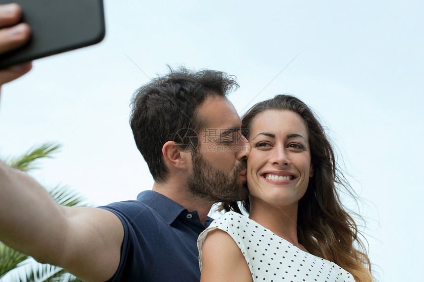 年轻男人亲吻女友的脸颊并使用手机自拍图片