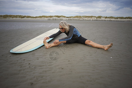 坐在沙滩上伸展冲浪板旁边的中老年女人图片