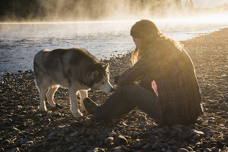 深沉沙哑的河岸上与狗狗玩耍的年轻女子背景