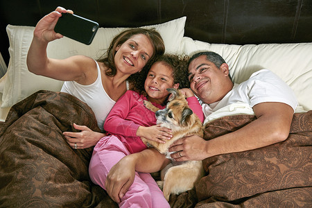 女孩与父母和狗在床上用手机自拍图片