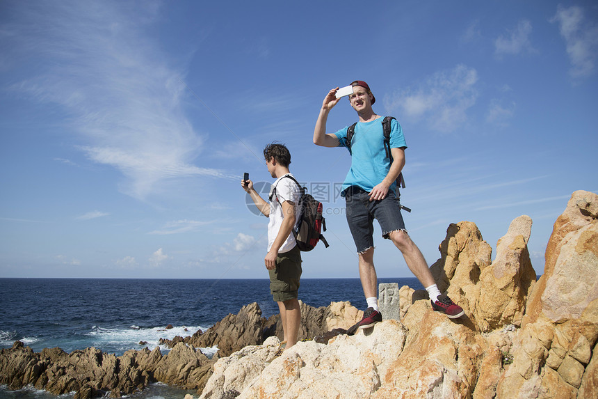青年男子站在岩石上使用智能手机拍照图片