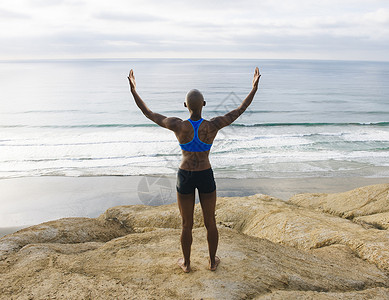 年轻女子站在海边的岩石上举起手臂的背影图片