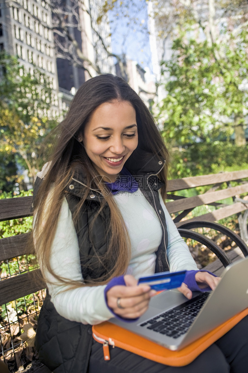 使用手提笔记本电脑信用卡坐在公园长椅上的年轻妇女图片