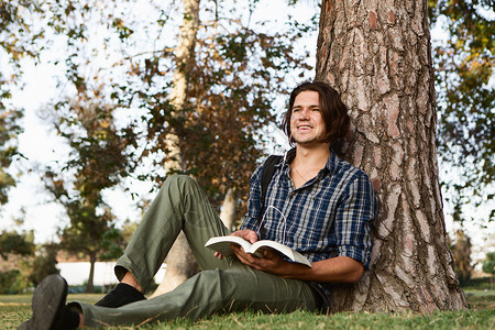 年轻人坐在树边拿着书微笑地看图片