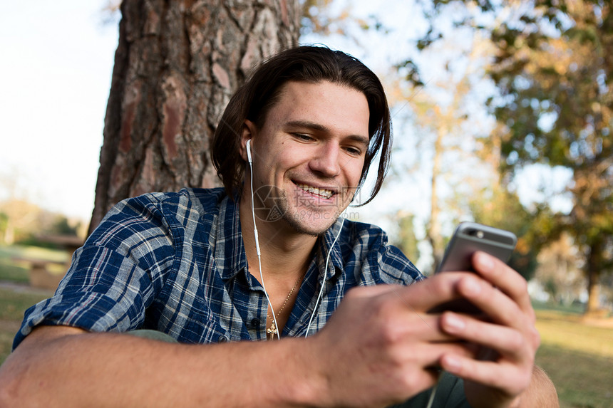 坐在树边的年轻人戴着耳机笑看智能手机图片