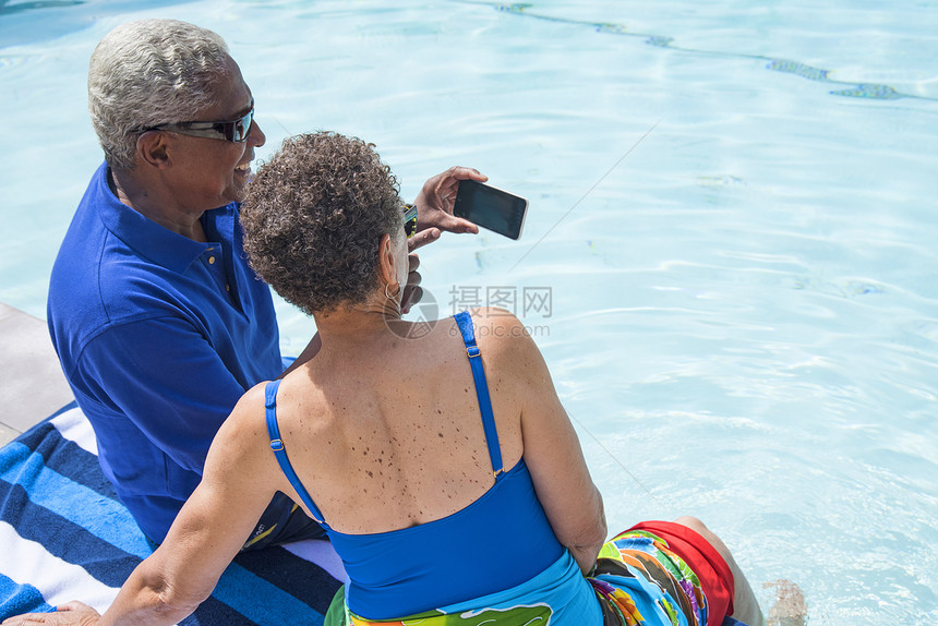 坐在游泳池旁边使用智能手机自拍的老年夫妇图片