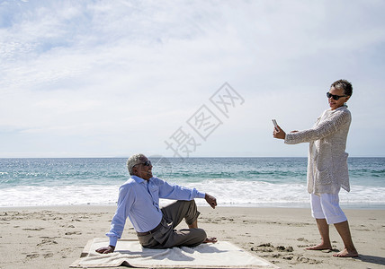 在海滩上的用智能手机拍摄丈夫照片的妻子图片