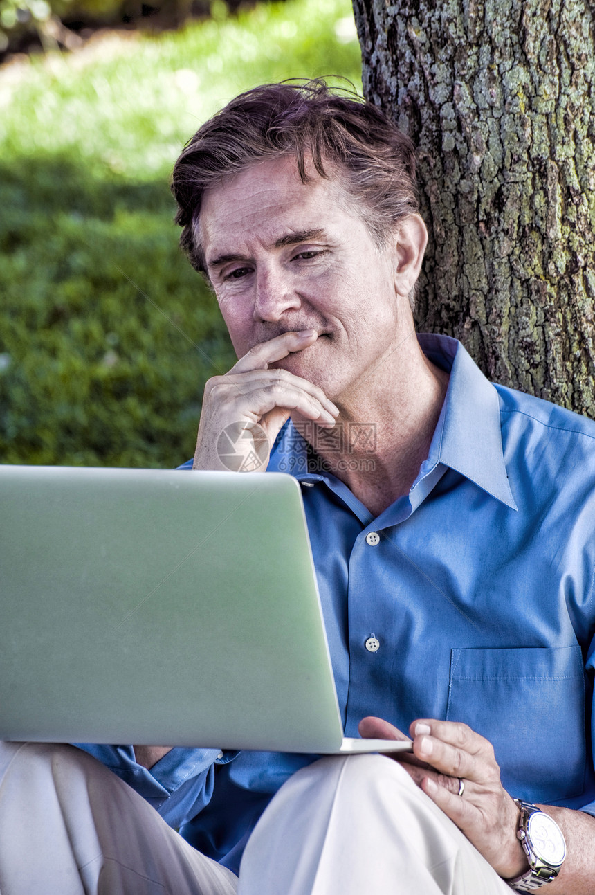 商人坐在户外靠在树上使用笔记本电脑图片