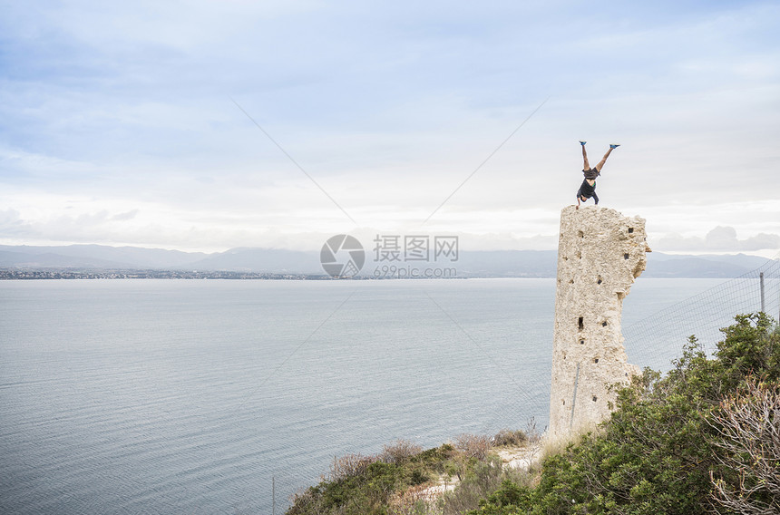 意大利卡亚里海岸被摧毁的塔台顶上做倒立的男攀岩者图片