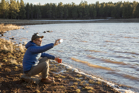 美国亚利桑那州Flagstaff中年男子坐在湖边用智能手机拍照图片