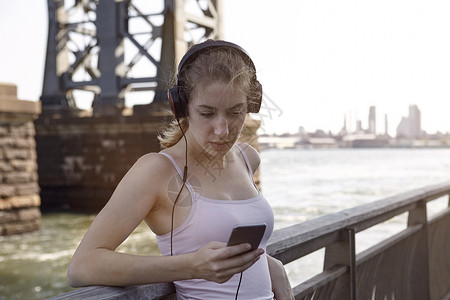 美国纽约州市江边桥上身戴耳机使用智能手的女性图片