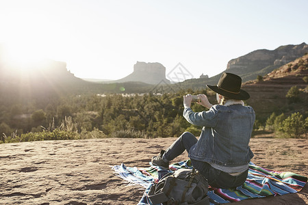美国亚利桑那州塞多纳一名妇女坐在沙漠中的毯子上用智能手机拍摄风景图片
