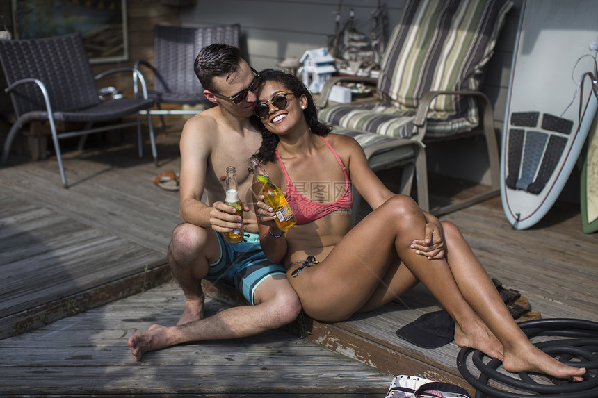 穿比基尼和泳裤的年轻夫妇在美国纽约州洛克威海滩的门廊上喝着啤酒图片